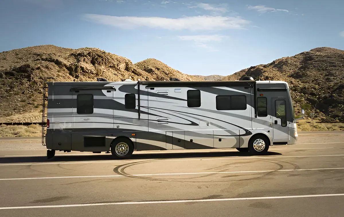 A gray and white recreational vehicle parked in a paved lot in front of rocky, desert hills under a sunny sky.