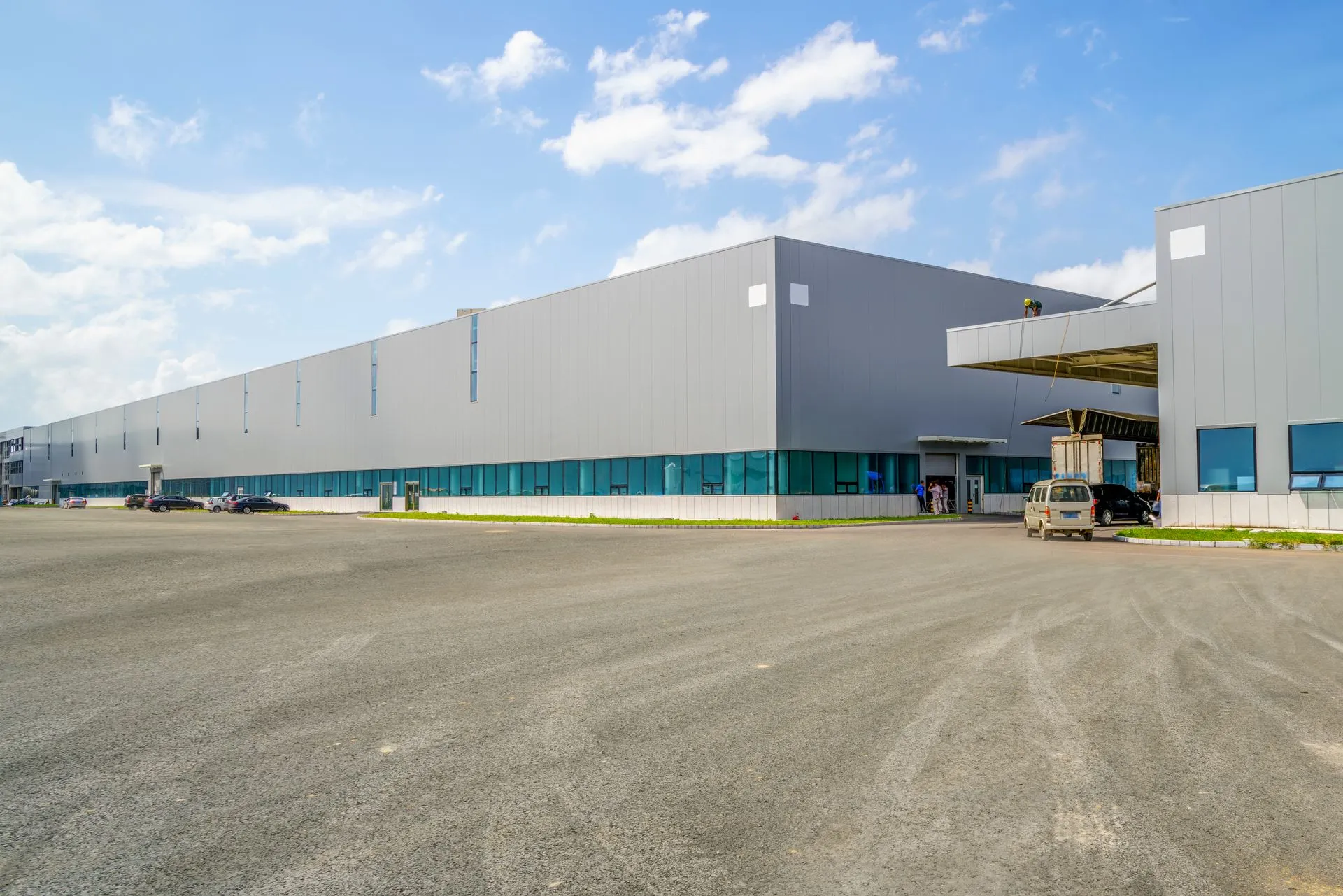 A long, gray, modern industrial warehouse building with a paved parking lot under a blue sky.